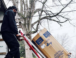 Delivery person using red hand truck to move large cardboard box near tree